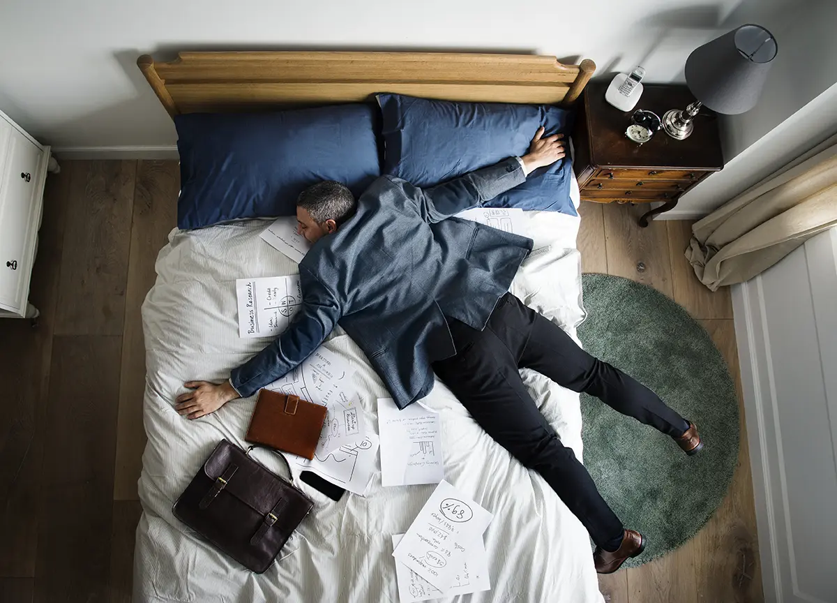 In Kleidung mit Arbeitstasche und Notizen ins Bett fallen, weil die Arbeit sich leer, erschöpfend oder auslaugend anfühlt, führt zu inneren Konflikten – reflektiert im Beziehungsworkshop 1030 Wien.
